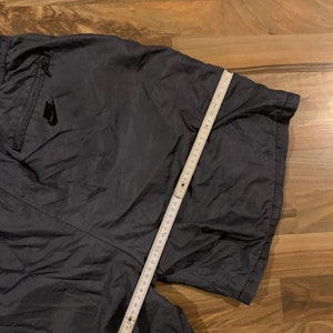 May include: Dark gray athletic shorts with a zippered pocket and a white Nike logo on the left side. The shorts are laid flat on a wooden surface with a measuring tape.