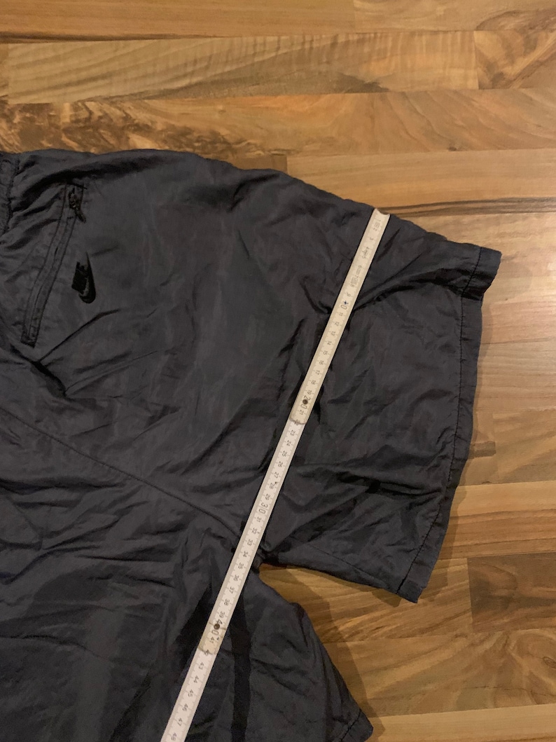May include: Dark gray athletic shorts with a zippered pocket and a white Nike logo on the left side. The shorts are laid flat on a wooden surface with a measuring tape.
