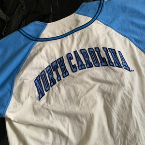 May include: White baseball jersey with blue sleeves and the text "NORTH CAROLINA" in blue embroidery on the back.