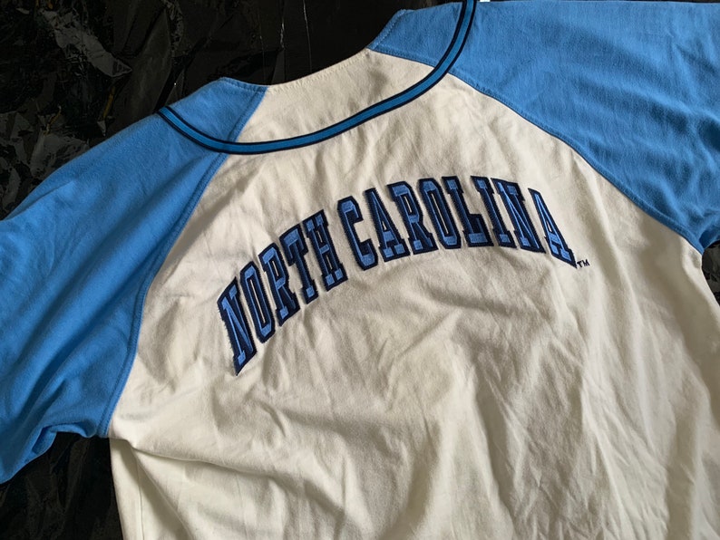 May include: White baseball jersey with blue sleeves and the text "NORTH CAROLINA" in blue embroidery on the back.