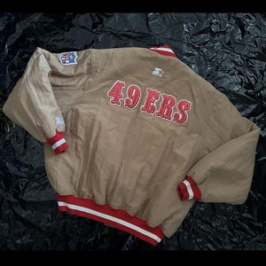 Könnte beinhalten: Beige Starter-Jacke mit roten und weißen Akzenten. Auf der Rückseite der Jacke steht in Rot der Schriftzug "49ERS" und das NFL-Logo. Die Ärmel und der Saum sind mit rot-weißen Streifen versehen. Ein Vintage-Sportbekleidungsstück.