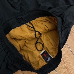 K&ouml;nnte beinhalten: Schwarze Sporthose mit goldfarbenem Futter und Kordelzug. Die Hose hat einen elastischen Bund und ein Etikett mit den Nike- und Jordan-Logos.