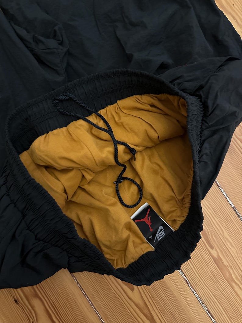 K&ouml;nnte beinhalten: Schwarze Sporthose mit goldfarbenem Futter und Kordelzug. Die Hose hat einen elastischen Bund und ein Etikett mit den Nike- und Jordan-Logos.