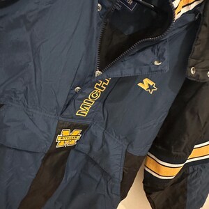 May include: A navy blue Starter jacket with black and yellow accents. The jacket features the word "MICHIGAN" in yellow, along with a yellow "M" logo. The jacket has a zipper and snap closures.