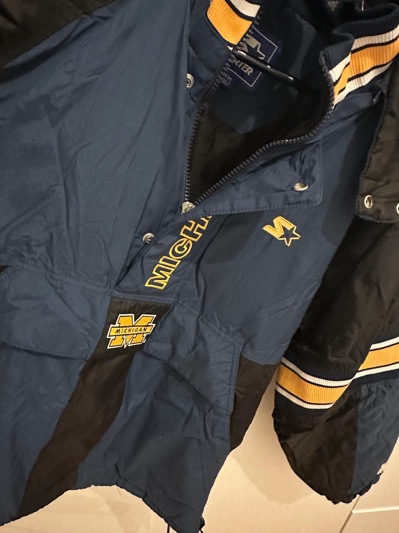 May include: A navy blue Starter jacket with black and yellow accents. The jacket features the word "MICHIGAN" in yellow, along with a yellow "M" logo. The jacket has a zipper and snap closures.