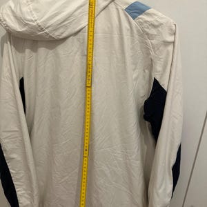Puede incluir: Chaqueta con capucha color crema con detalles en azul marino y azul claro. La chaqueta tiene una cremallera completa y una capucha. Una cinta m&eacute;trica amarilla se coloca verticalmente a lo largo de la chaqueta, midiendo unos 152 cm.