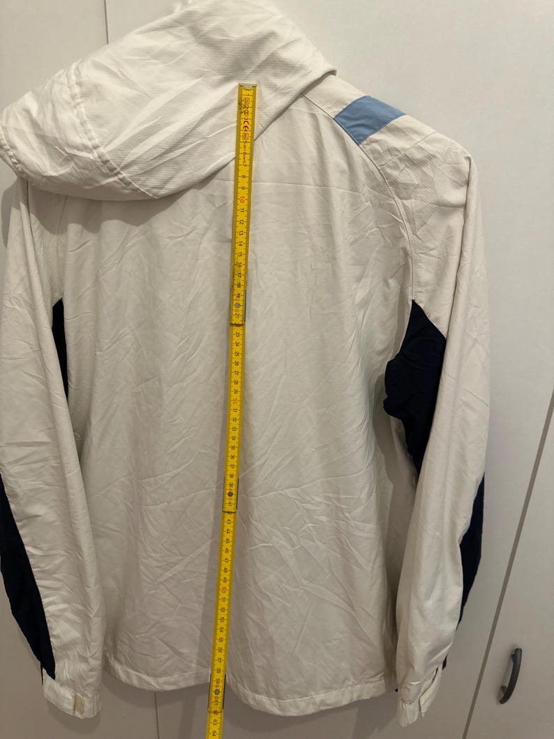 Puede incluir: Chaqueta con capucha color crema con detalles en azul marino y azul claro. La chaqueta tiene una cremallera completa y una capucha. Una cinta m&eacute;trica amarilla se coloca verticalmente a lo largo de la chaqueta, midiendo unos 152 cm.