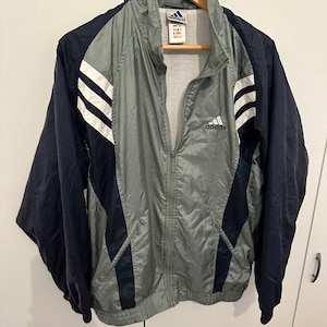 May include: A grey and navy blue windbreaker jacket with white stripes and the Adidas logo on the front.