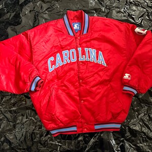 May include: Red satin bomber jacket with blue and white striped cuffs and collar. The jacket has a snap closure and features the word "CAROLINA" in blue lettering on the front.
