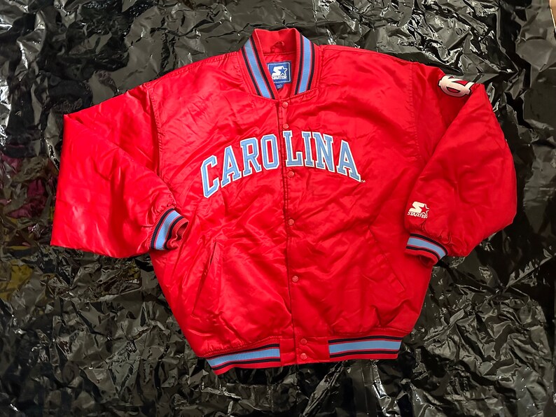 May include: Red satin bomber jacket with blue and white striped cuffs and collar. The jacket has a snap closure and features the word "CAROLINA" in blue lettering on the front.
