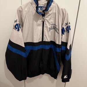 Könnte beinhalten: Eine graue, schwarze und blaue Orlando Magic Pullover-Jacke mit halbem Reißverschluss, hohem Kragen und dem auf die Brust gestickten Teamnamen. Die Ärmel zeigen den Teamnamen und ein Sternlogo, am linken Ärmelbündchen befindet sich ein Starter-Logo.