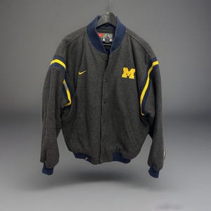 Könnte beinhalten: Graue Collegejacke mit Knöpfen und marineblauen und gelben Akzenten. Die Jacke hat den gelben Buchstaben "M" auf der Brust und ein Nike-Logo. Die Jacke hängt an einem schwarzen Kleiderbügel.