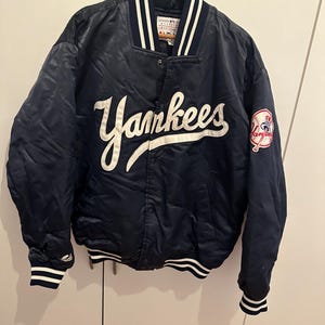 Può includere: Giacca da baseball dei Yankees in raso blu navy con scritte e finiture bianche. La giacca presenta la scritta "Yankees" sul petto e una toppa con il logo della squadra sulla manica. Il colletto e i polsini hanno strisce bianche.