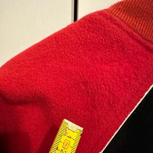May include: Close-up of a red fabric with a textured surface. The fabric is trimmed with a white border and black fabric. A yellow ruler is placed on the fabric, showing measurements in inches.