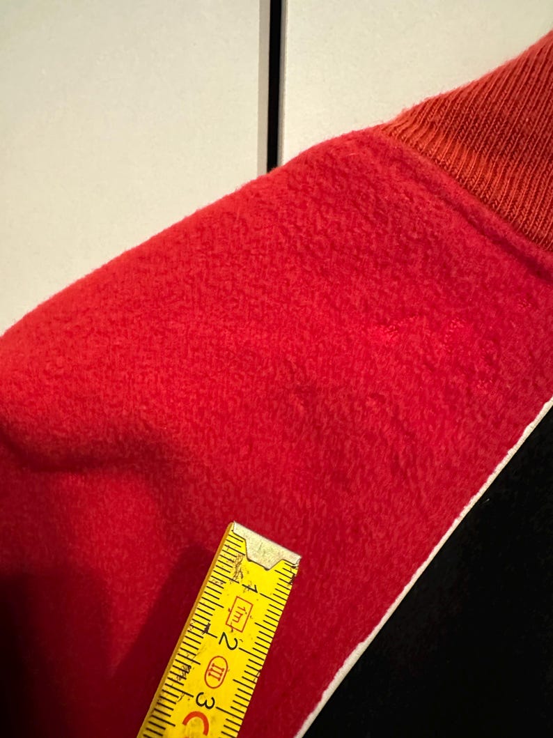 May include: Close-up of a red fabric with a textured surface. The fabric is trimmed with a white border and black fabric. A yellow ruler is placed on the fabric, showing measurements in inches.