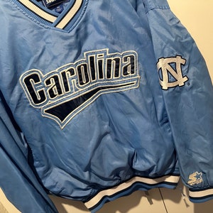 K&ouml;nnte beinhalten: Eine hellblaue Windbreaker-Jacke mit einem wei&szlig;-blau gestreiften Kragen. Die Jacke hat ein gro&szlig;es "Carolina"-Logo auf der Vorderseite und ein kleineres "N"-Logo auf dem &Auml;rmel. Die Jacke ist von Starter.