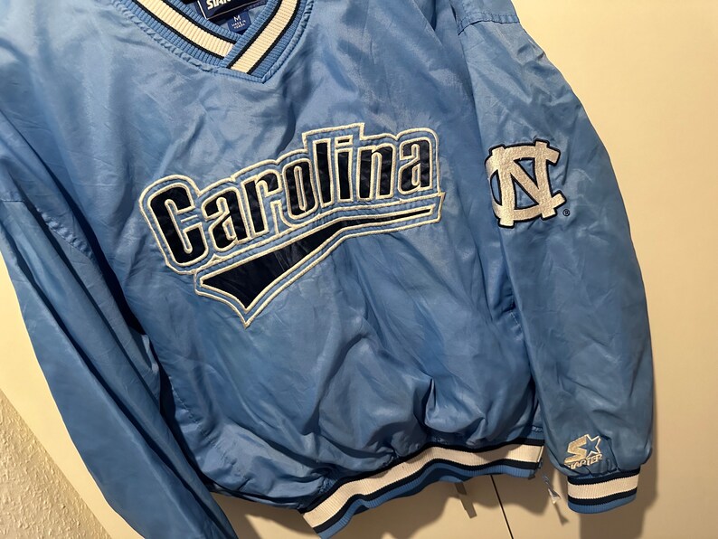 K&ouml;nnte beinhalten: Eine hellblaue Windbreaker-Jacke mit einem wei&szlig;-blau gestreiften Kragen. Die Jacke hat ein gro&szlig;es "Carolina"-Logo auf der Vorderseite und ein kleineres "N"-Logo auf dem &Auml;rmel. Die Jacke ist von Starter.