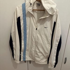 Puede incluir: Chaqueta cortavientos Nike blanca con capucha, una franja azul claro en el lado izquierdo y detalles en azul marino en las mangas. Presenta un logotipo de Nike negro en el pecho. La chaqueta est&aacute; colgada en una percha de madera.