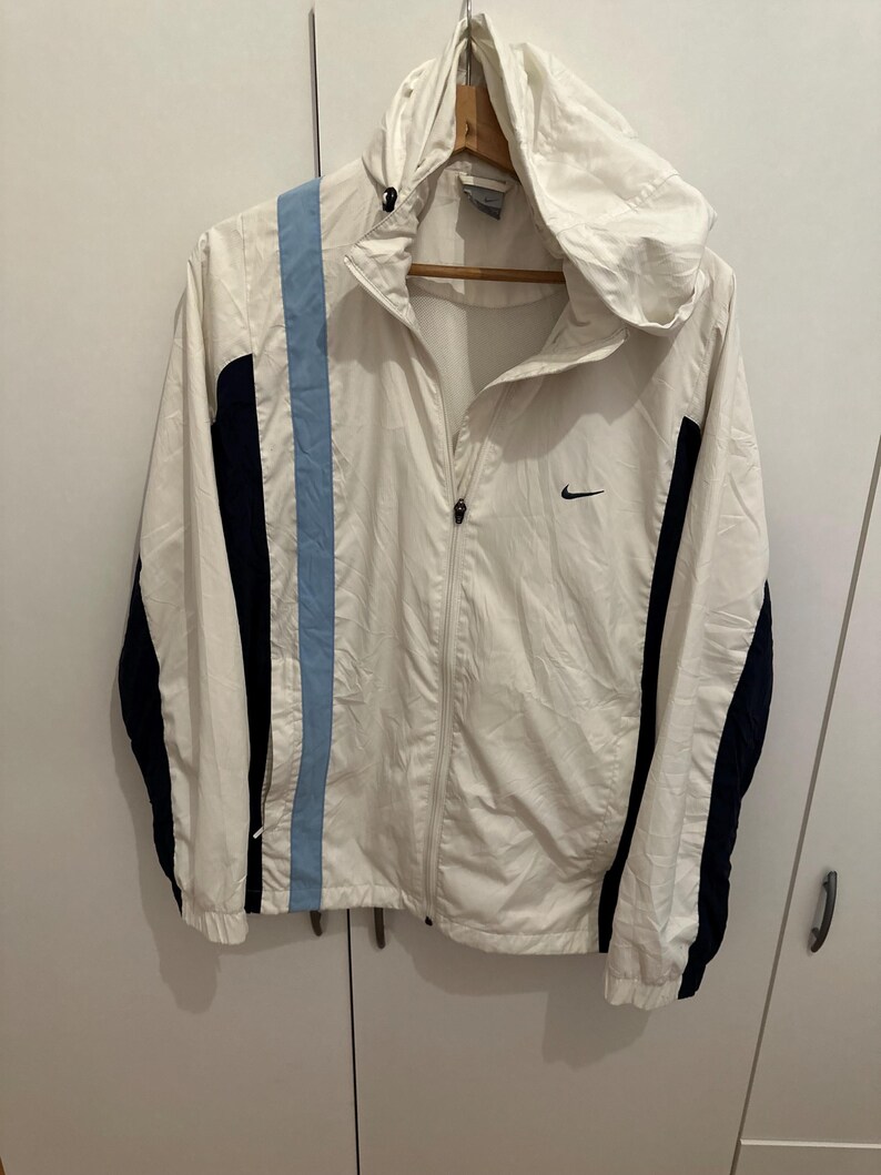 Puede incluir: Chaqueta cortavientos Nike blanca con capucha, una franja azul claro en el lado izquierdo y detalles en azul marino en las mangas. Presenta un logotipo de Nike negro en el pecho. La chaqueta est&aacute; colgada en una percha de madera.