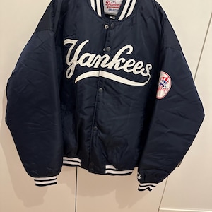 Op de afbeelding: Marineblauwe Yankees honkbaljack met witte letters en een teamlogopatch. De jas heeft een knoopsluiting, een gestreepte kraag en gestreepte manchetten en zoom. Het Starter-logo is zichtbaar op de kraag.