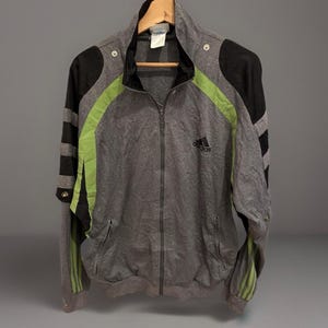 May include: A gray Adidas track jacket with black and lime green accents. The jacket features a full zipper, side pockets, and the Adidas logo on the chest. The sleeves have black stripes and the jacket is hanging on a wooden hanger.