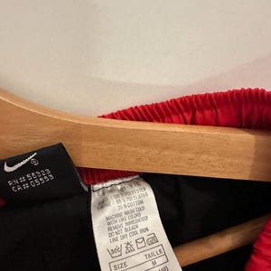 May include: A black and red tag with white text. The tag has the following information: RN #55323 CA#05553 54-09-01-VTG 100% POLYESTER LINING: 65% POLYESTER 35% COTTON MACHINE WASH COLD WITH LIKE COLORS REMOVE IMMEDIATELY DO NOT BLEACH LINE DRY COOL IRON 30C SIZE M TAILLE M MADE IN THAILAND RN #56323