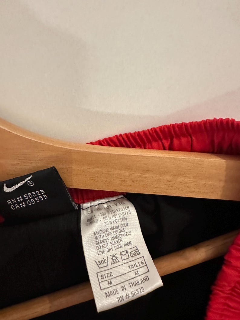 May include: A black and red tag with white text. The tag has the following information: RN #55323 CA#05553 54-09-01-VTG 100% POLYESTER LINING: 65% POLYESTER 35% COTTON MACHINE WASH COLD WITH LIKE COLORS REMOVE IMMEDIATELY DO NOT BLEACH LINE DRY COOL IRON 30C SIZE M TAILLE M MADE IN THAILAND RN #56323