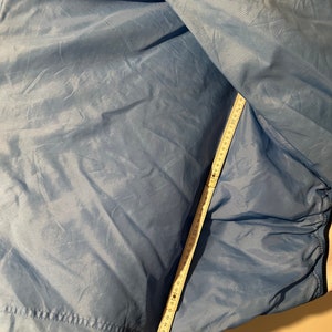 K&ouml;nnte beinhalten: Eine blaue Nylonjacke mit einem wei&szlig;en Rei&szlig;verschluss und wei&szlig;en Bes&auml;tzen. Die Jacke liegt flach und ein Ma&szlig;band ist an der Seite der Jacke angebracht.