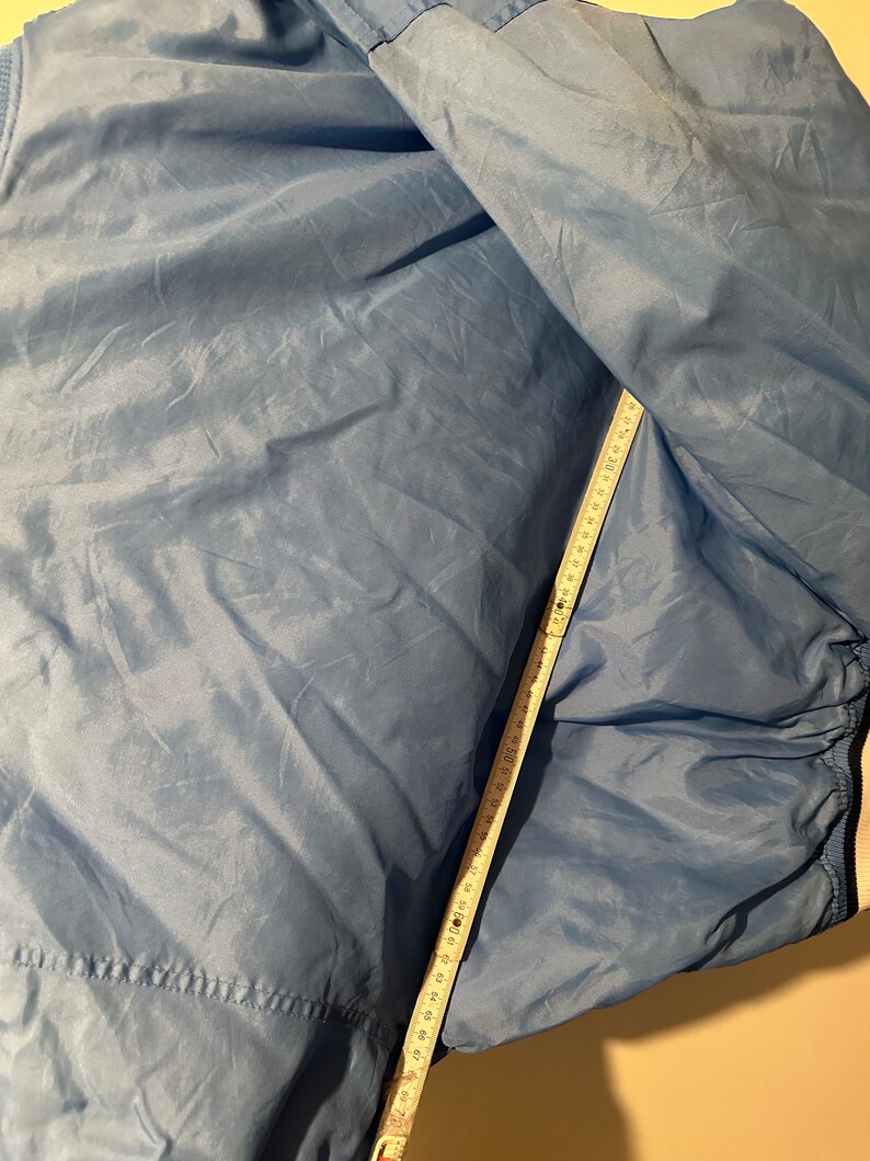 K&ouml;nnte beinhalten: Eine blaue Nylonjacke mit einem wei&szlig;en Rei&szlig;verschluss und wei&szlig;en Bes&auml;tzen. Die Jacke liegt flach und ein Ma&szlig;band ist an der Seite der Jacke angebracht.