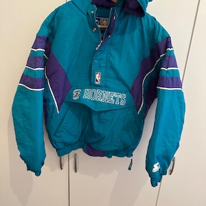 May include: A teal and purple Charlotte Hornets pullover jacket with a hood, a front pocket, and a partial zip closure. The jacket features the NBA logo on the chest, the Hornets team name in white text, and a Starter logo on the left sleeve cuff.