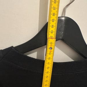 May include: A black crew neck sweatshirt hanging on a black hanger. A yellow ruler is placed next to the sweatshirt, showing measurements in centimetres. The ruler is marked from 68 to 90 cm.