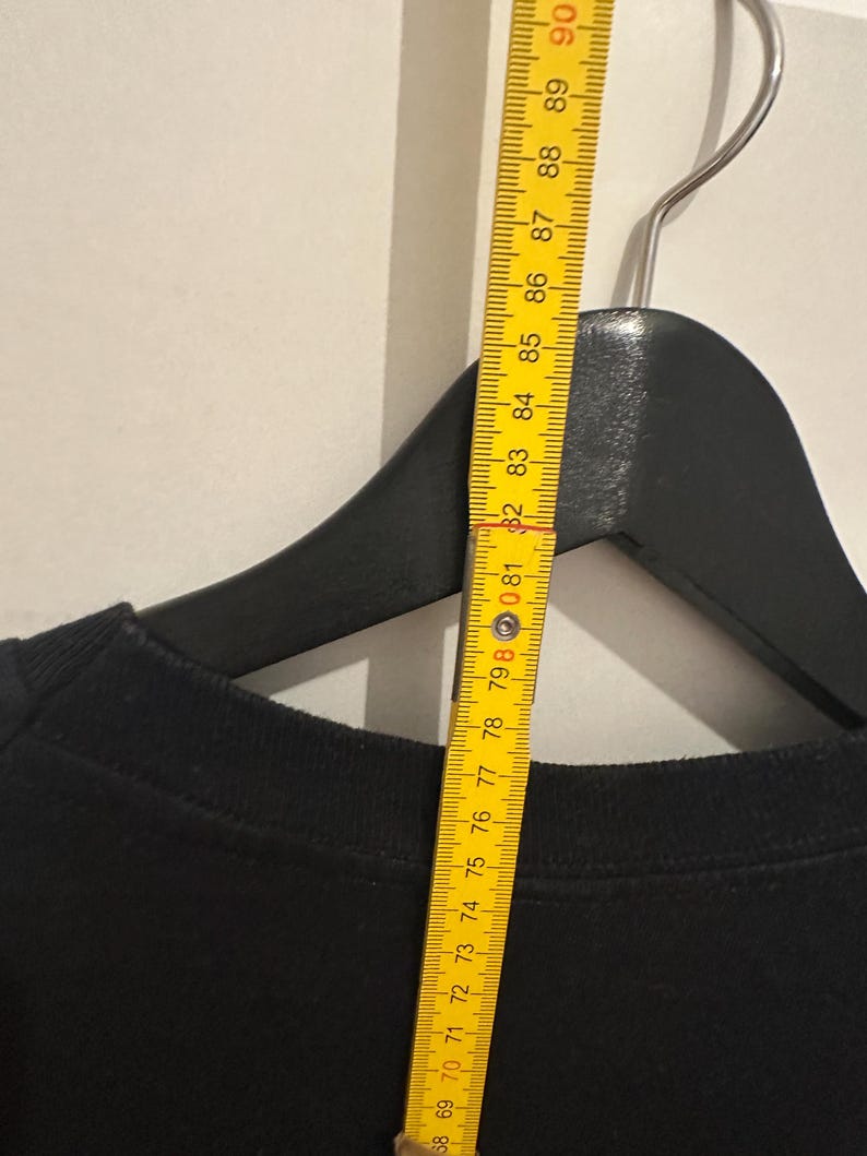 May include: A black crew neck sweatshirt hanging on a black hanger. A yellow ruler is placed next to the sweatshirt, showing measurements in centimetres. The ruler is marked from 68 to 90 cm.