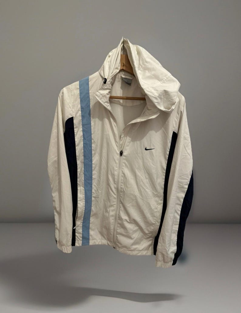 Puede incluir: Chaqueta con capucha Nike blanca con cremallera completa. La chaqueta presenta una franja azul claro en el lado izquierdo y paneles azul marino en las mangas. Peque&ntilde;o logotipo negro de Nike en el pecho.