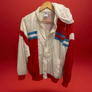 May include: A vintage white, red, and blue athletic jacket with a hood, hanging on a wooden hanger. The jacket features red panels on the sleeves and shoulders, with blue stripes. The jacket has a mesh lining and a zipper closure.