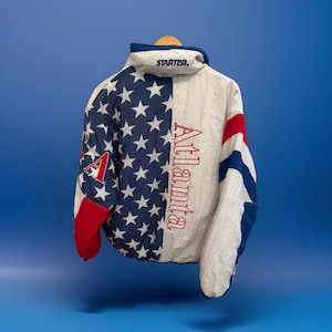 Könnte beinhalten: Vintage Starter Jacke mit patriotischem Design. Die linke Seite zeigt ein blaues Feld mit weißen Sternen, während die rechte Seite weiß mit roten und blauen Akzenten ist. Das Wort "Atlanta" ist vertikal in Rot gedruckt.