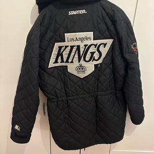 Könnte beinhalten: Schwarze, gesteppte Jacke mit Kapuze, mit dem Logo "STARTER" und dem Text "Los Angeles KINGS" in Weiß. Die Jacke hat einen Kordelzug in der Taille und einen Teamlogo-Aufnäher am Ärmel.
