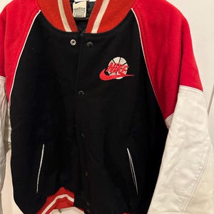 May include: Black, red, and white varsity jacket with a basketball logo. The jacket features a black body, red sleeves and collar, and white accents. The Nike logo is embroidered on the chest.
