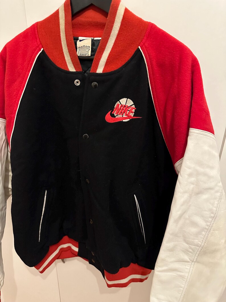 May include: Black, red, and white varsity jacket with a basketball logo. The jacket features a black body, red sleeves and collar, and white accents. The Nike logo is embroidered on the chest.