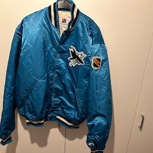 Könnte beinhalten: Eine blaue Satin-Bomberjacke mit einem weißen und schwarzen gestreiften Kragen und Bündchen. Die Jacke hat ein San Jose Sharks-Logo auf der linken Brust und ein NHL-Logo auf dem rechten Ärmel.