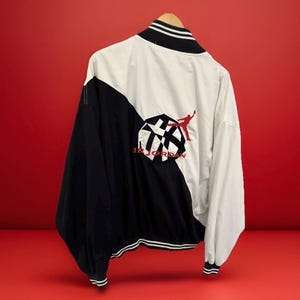 Könnte beinhalten: Schwarz-weiße Jacke mit rotem Logo und Text. Die Jacke hat einen schwarzen Kragen und Manschetten mit weißen Streifen. Auf der Rückseite der Jacke befindet sich eine große Grafik mit den Worten "IR JORDAN".