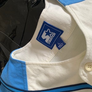 May include: A white baseball jersey with blue and navy blue stripes. The jersey has a blue Starter logo with a white star and the text "Starter" in white. The jersey has a size tag that says "L" and "Made in Korea".