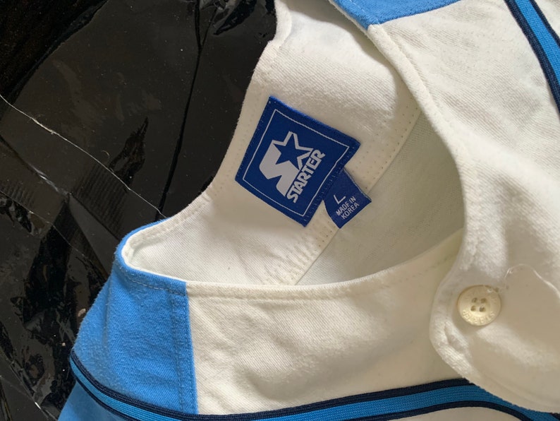 May include: A white baseball jersey with blue and navy blue stripes. The jersey has a blue Starter logo with a white star and the text "Starter" in white. The jersey has a size tag that says "L" and "Made in Korea".