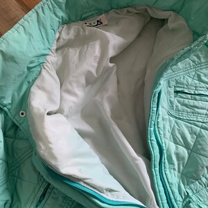 May include: A mint green quilted jacket with a zipper closure. The jacket has a white lining with a repeated pattern of a small square.