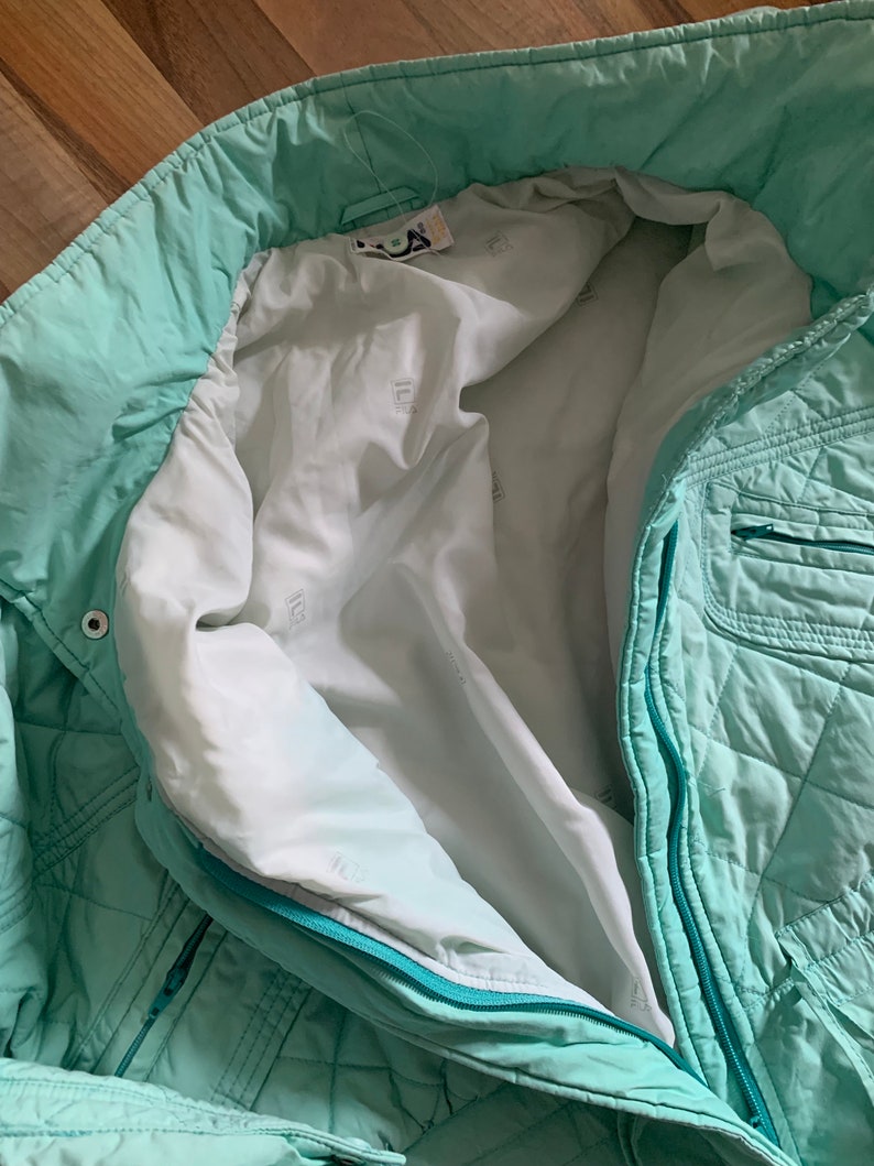 May include: A mint green quilted jacket with a zipper closure. The jacket has a white lining with a repeated pattern of a small square.