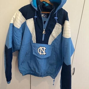 May include: A vintage Starter pullover jacket in light blue, navy, and white. The jacket features a hood, a front pocket with the University of North Carolina logo, and a half-zip closure. The sleeves are navy blue with the Starter logo.