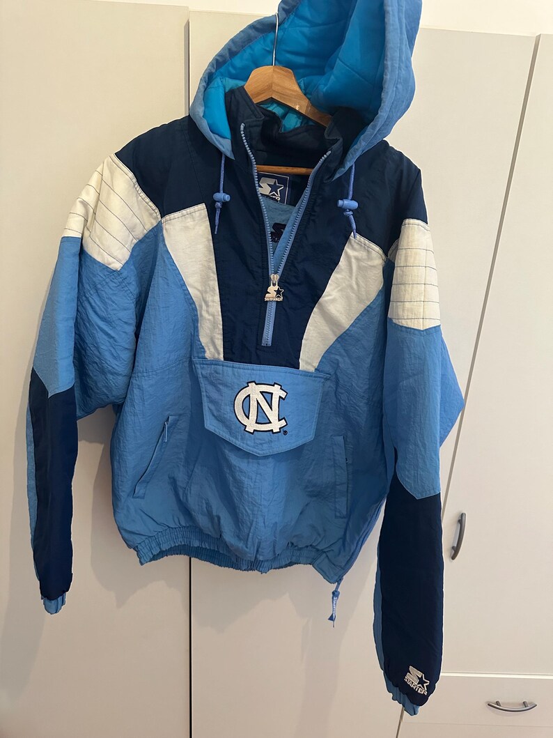 May include: A vintage Starter pullover jacket in light blue, navy, and white. The jacket features a hood, a front pocket with the University of North Carolina logo, and a half-zip closure. The sleeves are navy blue with the Starter logo.