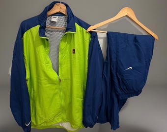 May include: A two-piece athletic tracksuit in a vibrant colour scheme. The jacket is lime green with navy blue sleeves and accents, featuring a zip and the Nike logo. The matching trousers are navy blue with a white Nike swoosh.