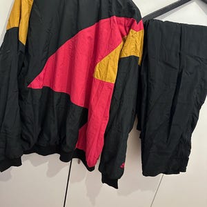 K&ouml;nnte beinhalten: Vintage Trainingsanzug in Schwarz, Pink und Gold. Die Jacke hat ein Colorblock-Design mit einem pinkfarbenen und goldenen Chevron-Muster auf der Brust. Die passende Hose ist einfarbig schwarz. Das Nike-Logo ist auf der Jacke sichtbar.