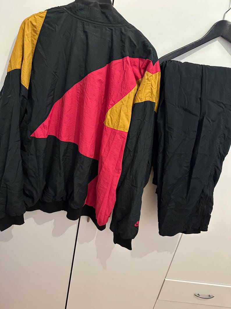 K&ouml;nnte beinhalten: Vintage Trainingsanzug in Schwarz, Pink und Gold. Die Jacke hat ein Colorblock-Design mit einem pinkfarbenen und goldenen Chevron-Muster auf der Brust. Die passende Hose ist einfarbig schwarz. Das Nike-Logo ist auf der Jacke sichtbar.
