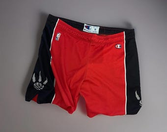 May include: Red basketball shorts with black side panels featuring a claw graphic and white accents. The shorts have a black waistband and the Champion logo. The NBA logo is also visible.
